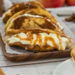 Three dessert tacos filled with cream and drizzled with caramel sauce are arranged on a wooden tray, with apples and cinnamon sticks in the background.