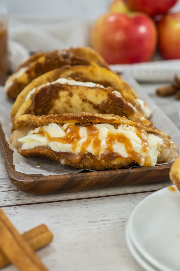 Three dessert tacos filled with cream and drizzled with caramel sauce are arranged on a wooden tray, with apples and cinnamon sticks in the background.
