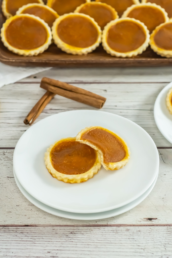 Two pumpkin tarts on a white plate, one cut in half, with a tray of more tarts and two cinnamon sticks in the background on a wooden surface.