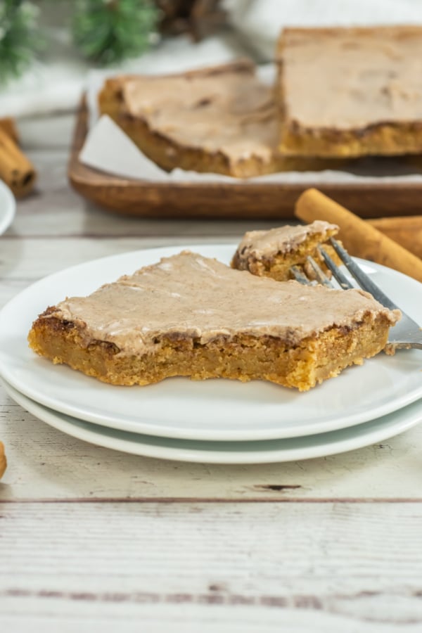 A square slice of frosted cookie bar with a bite taken out, served on a white plate with a fork. More cookie bars are visible in the background.
