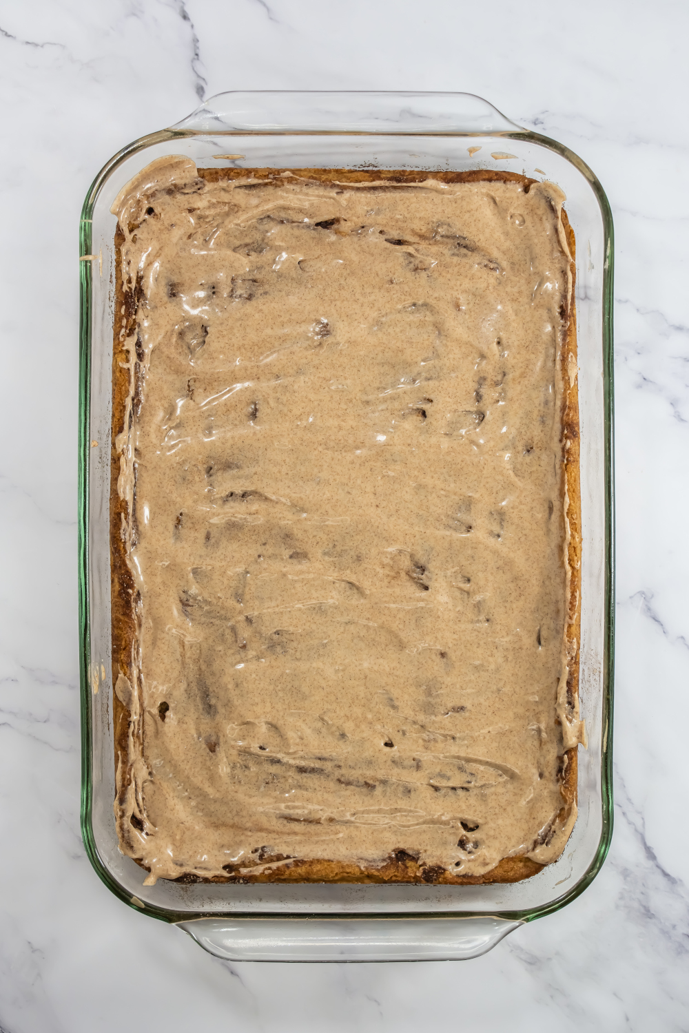 A rectangular glass baking dish filled with a baked dessert, topped with a layer of light brown frosting, on a marble surface.