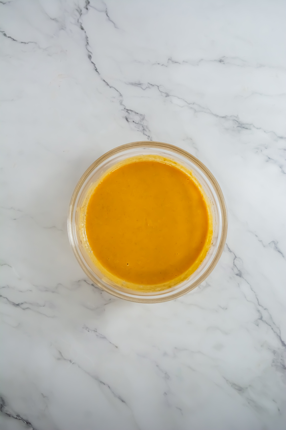 A glass bowl filled with orange-colored sauce or dressing sits on a white marble surface.