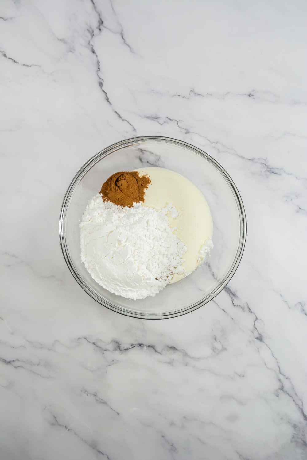 A glass bowl containing powdered sugar, ground cinnamon, and heavy cream on a white marble surface.
