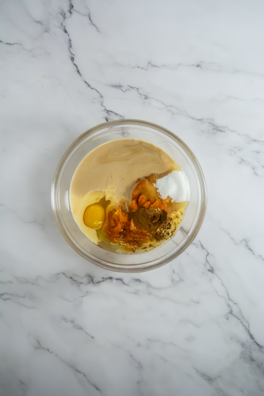 A glass bowl on a marble surface containing tahini, an egg, brown sugar, white sugar, baking soda, and vanilla extract, before mixing.