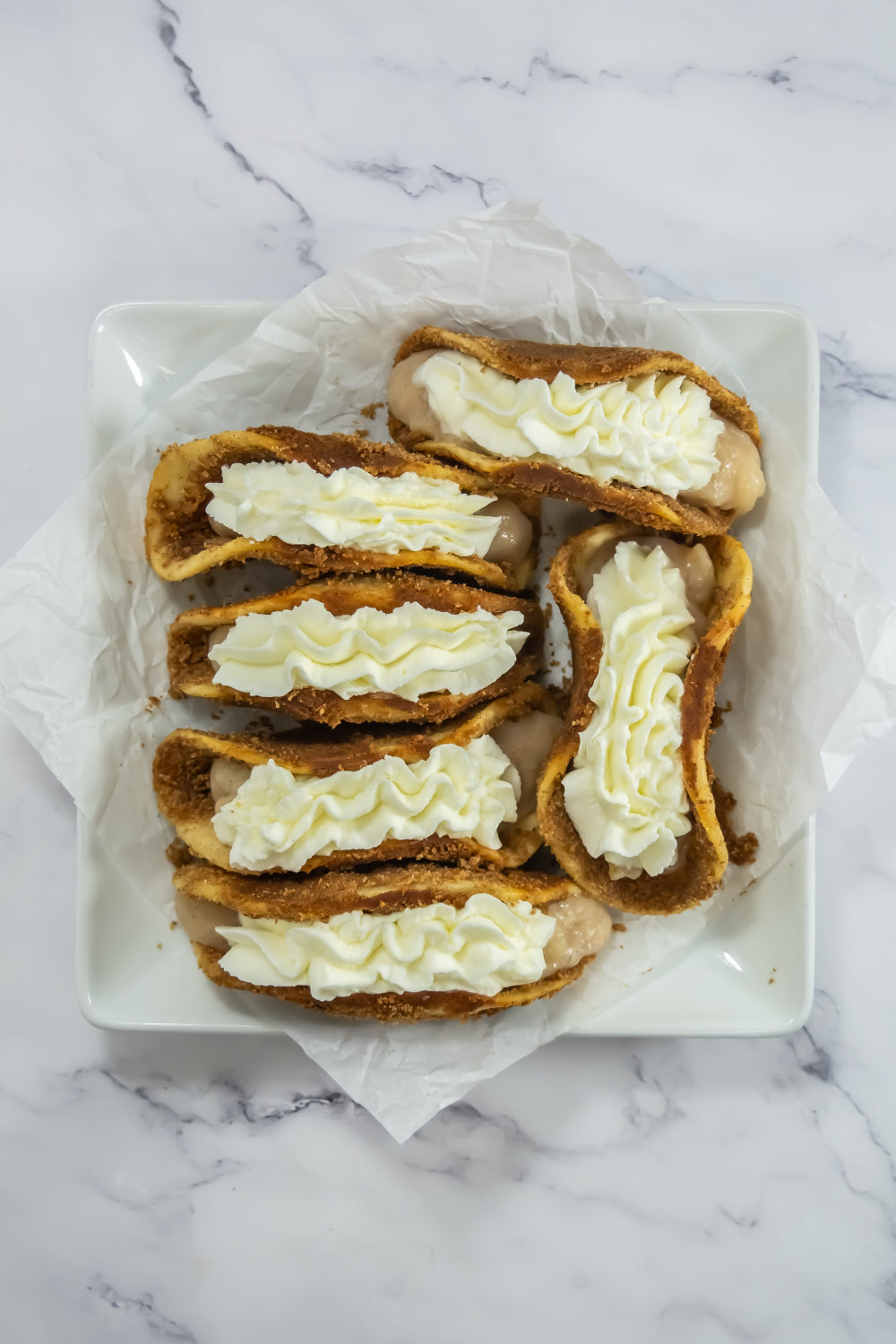 Six cannoli shells filled with cream are arranged on a square white plate lined with parchment paper, set on a marble surface.