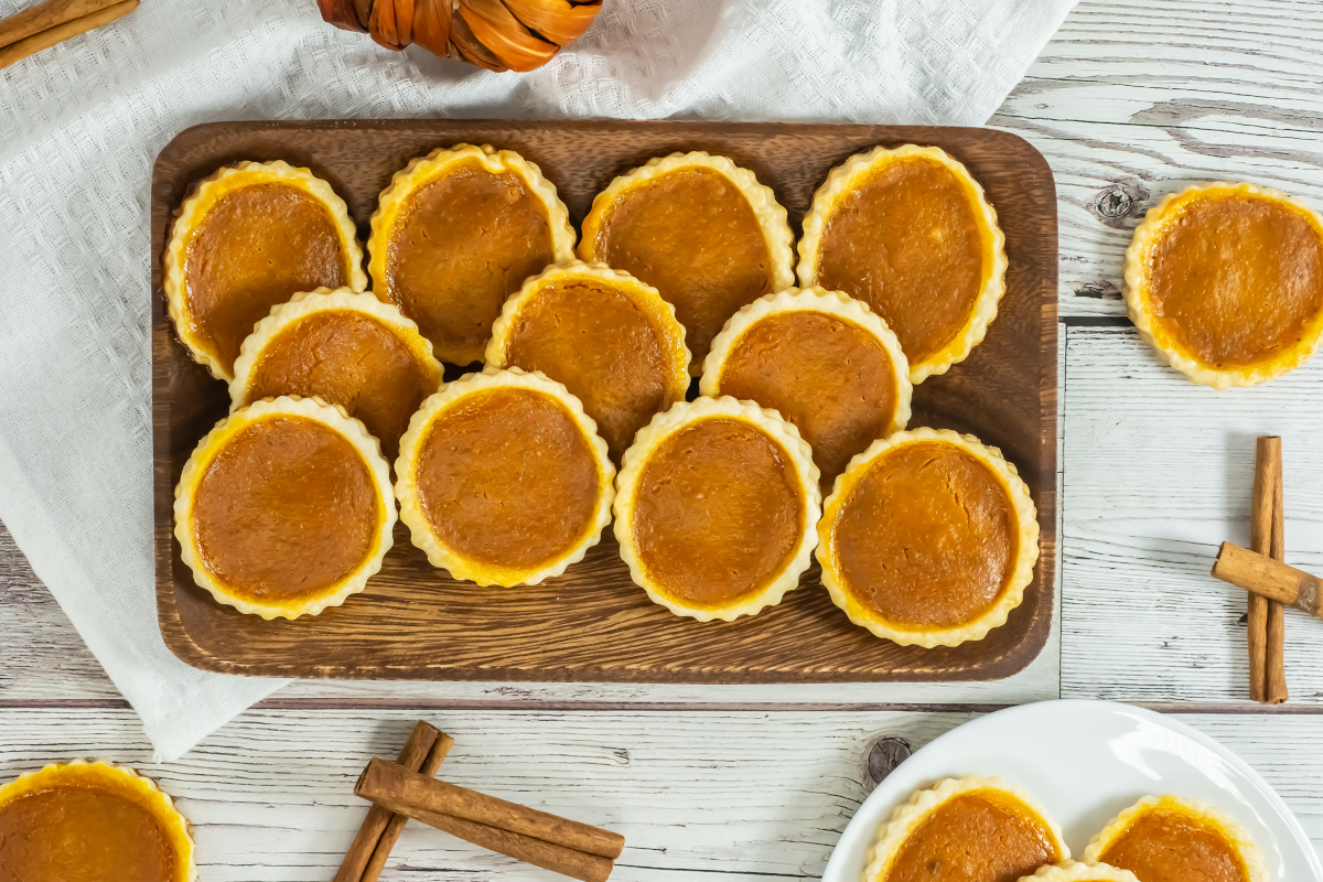 A wooden board topped with small pumpkin tarts, arranged neatly on a white table with cinnamon sticks and a white napkin nearby.