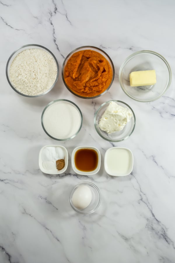 Nine baking ingredients are arranged on a marble surface, including flour, pumpkin puree, butter, sugar, cream cheese, baking powder, spices, vanilla extract, milk, and an egg.