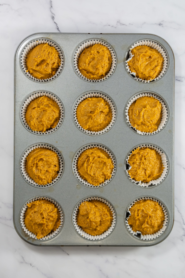 A muffin tin filled with twelve paper liners, each containing a portion of raw pumpkin muffin batter, on a marble surface.
