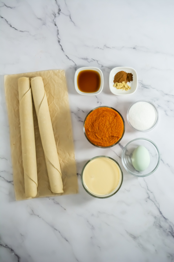 Two rolled pie crusts on parchment paper, with bowls of pumpkin puree, sugar, evaporated milk, an egg, spices, and vanilla extract arranged on a marble surface.