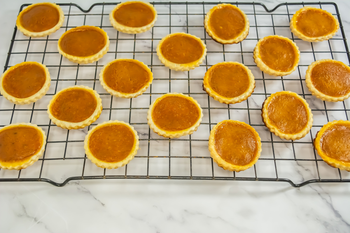 Mini pumpkin pies are cooling on a black wire rack placed on a white marble surface.