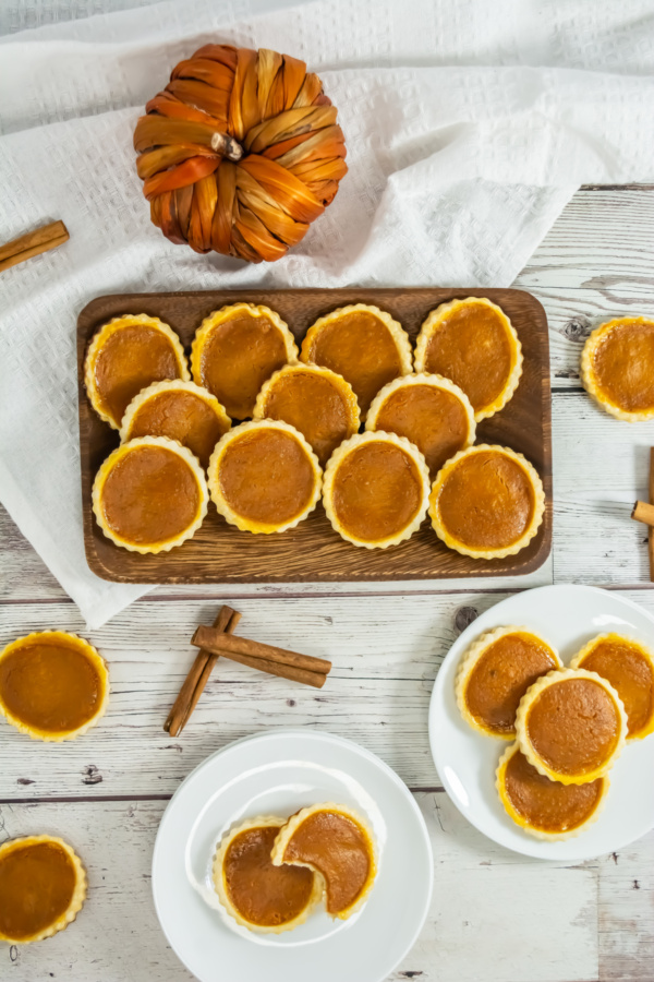Mini pumpkin pies arranged on a wooden tray and white plates, with cinnamon sticks and a decorative pumpkin on a rustic white table.