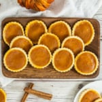 A wooden tray holds several pumpkin tarts on a white table, with cinnamon sticks and a plate of more tarts nearby.