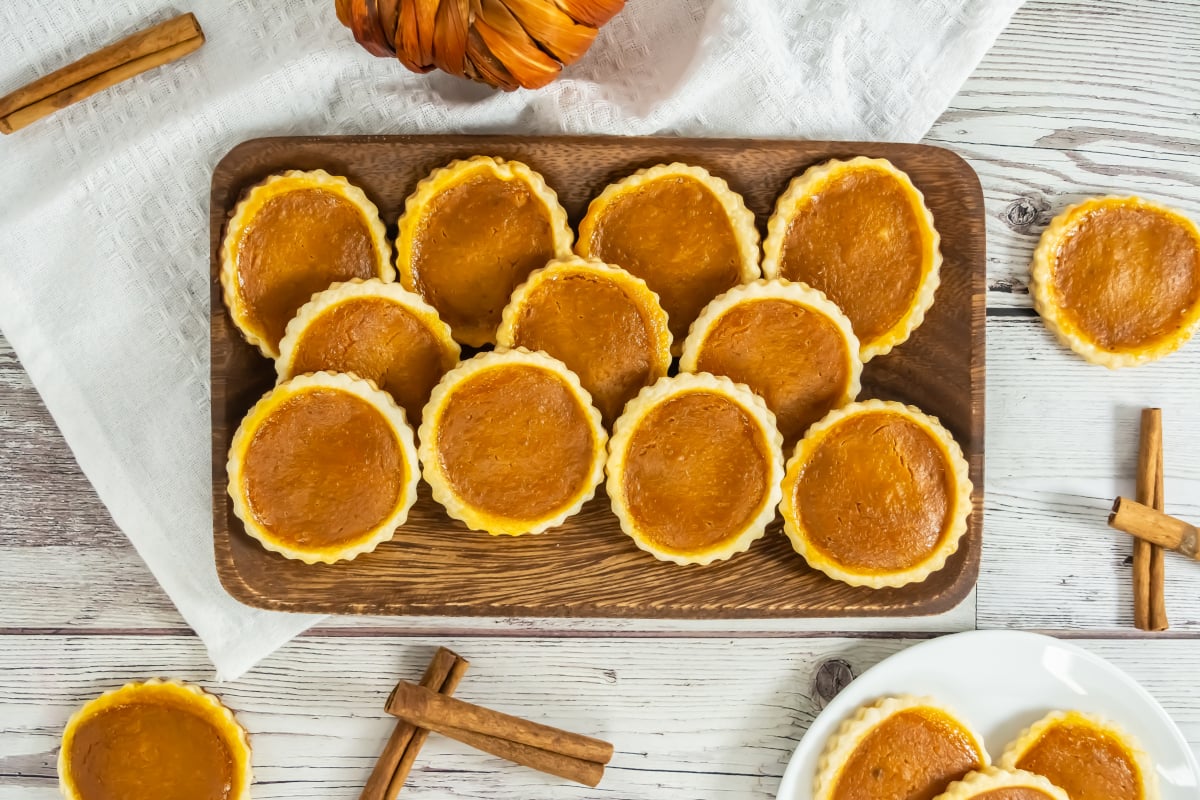 A wooden tray holds several pumpkin tarts on a white table, with cinnamon sticks and a plate of more tarts nearby.