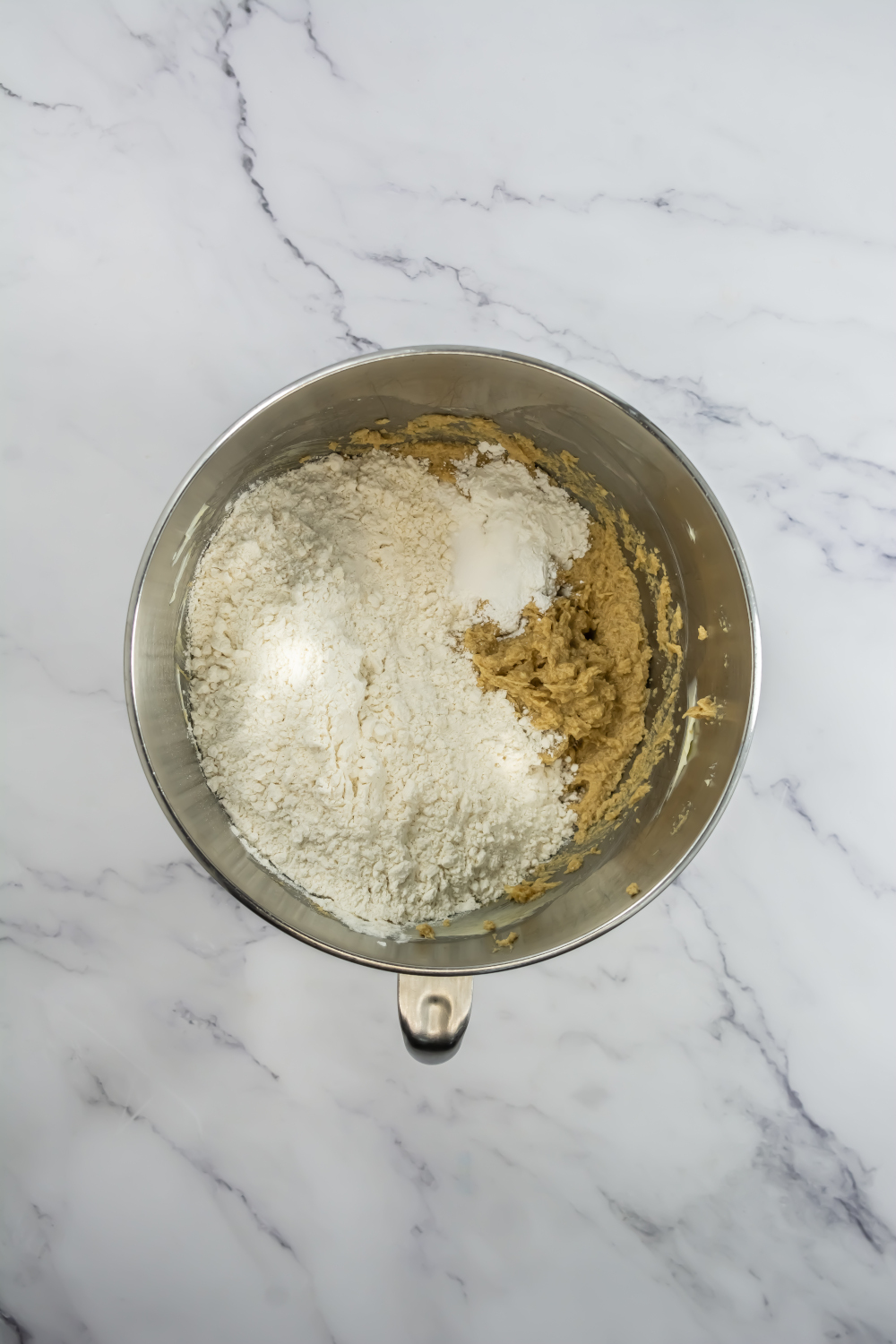 A metal mixing bowl containing flour, baking powder, and cookie dough mixture on a white marble surface.