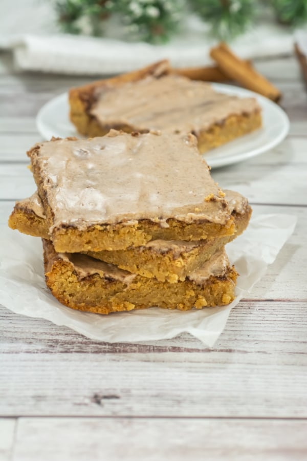 Three frosted cookie bars are stacked on parchment paper with more bars on a white plate in the background, set on a wooden surface.