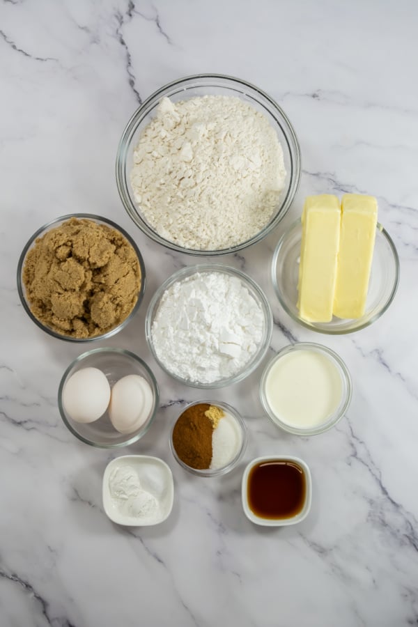 A top-down view of baking ingredients in bowls on a marble surface, including flour, brown sugar, powdered sugar, butter, eggs, milk, vanilla extract, baking powder, and spices.