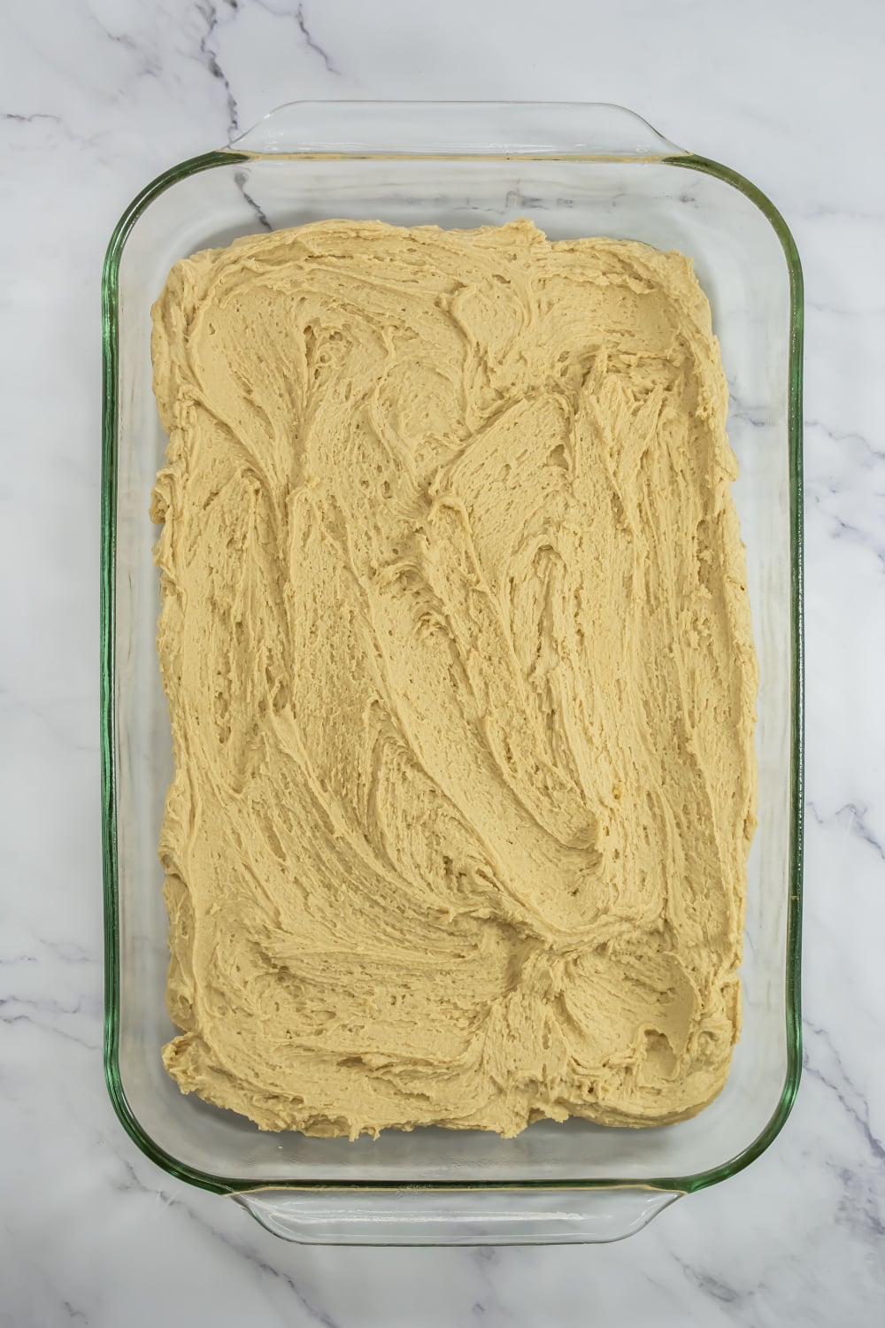 A glass baking dish filled with an even layer of unbaked light brown batter on a white marble surface.