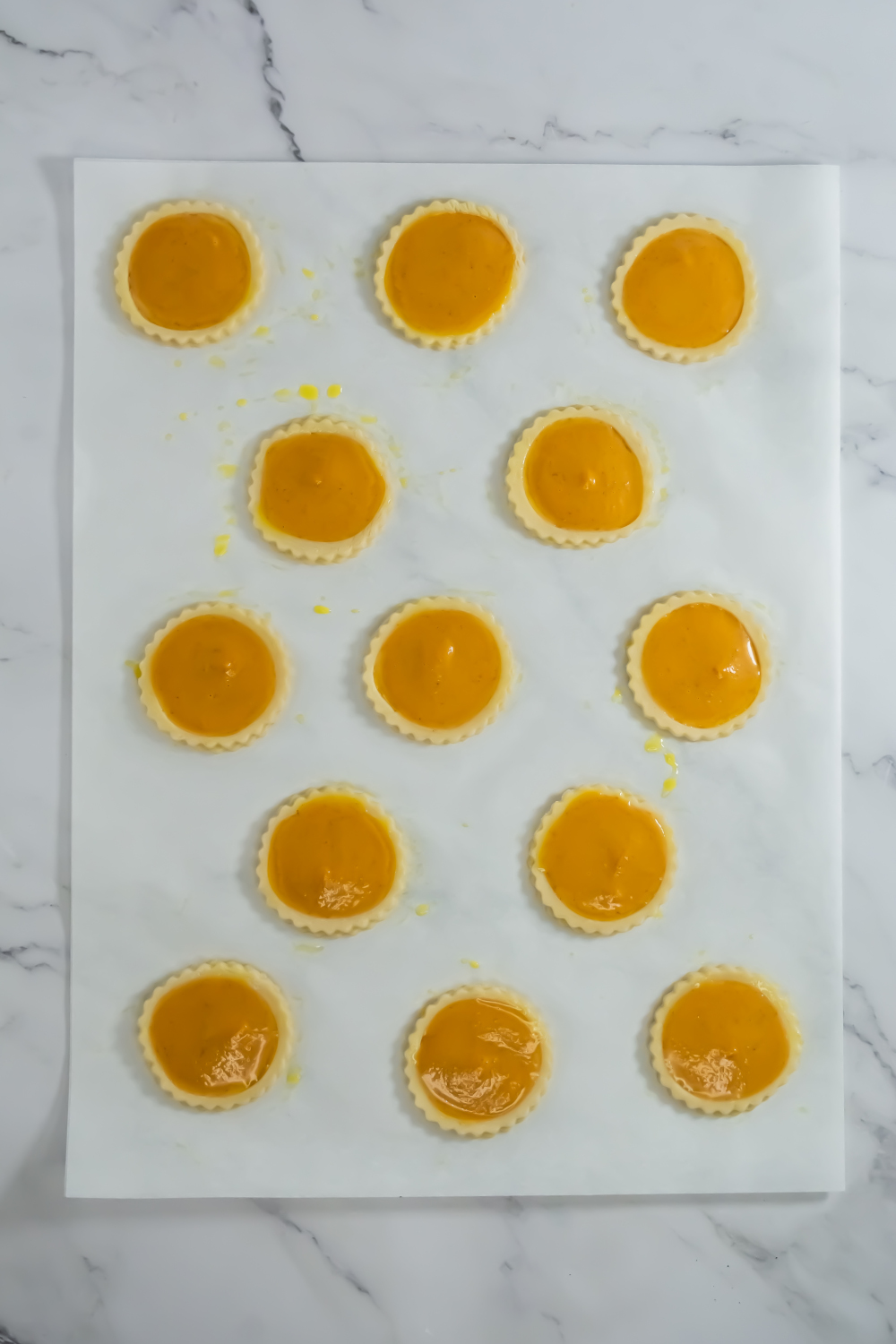 Fourteen unbaked custard tarts are arranged evenly on a sheet of parchment paper atop a marble surface.