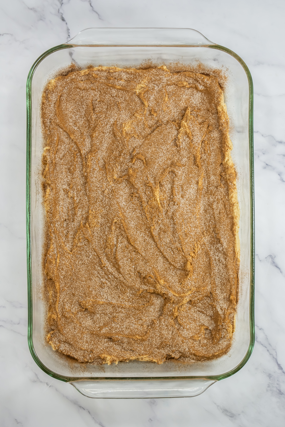 A glass baking dish filled with unbaked Snickerdoodle Cookie Bars Recipe batter, topped with cinnamon and sugar, rests on a marble surface—perfect for easy dessert bars at bake sales or holidays.