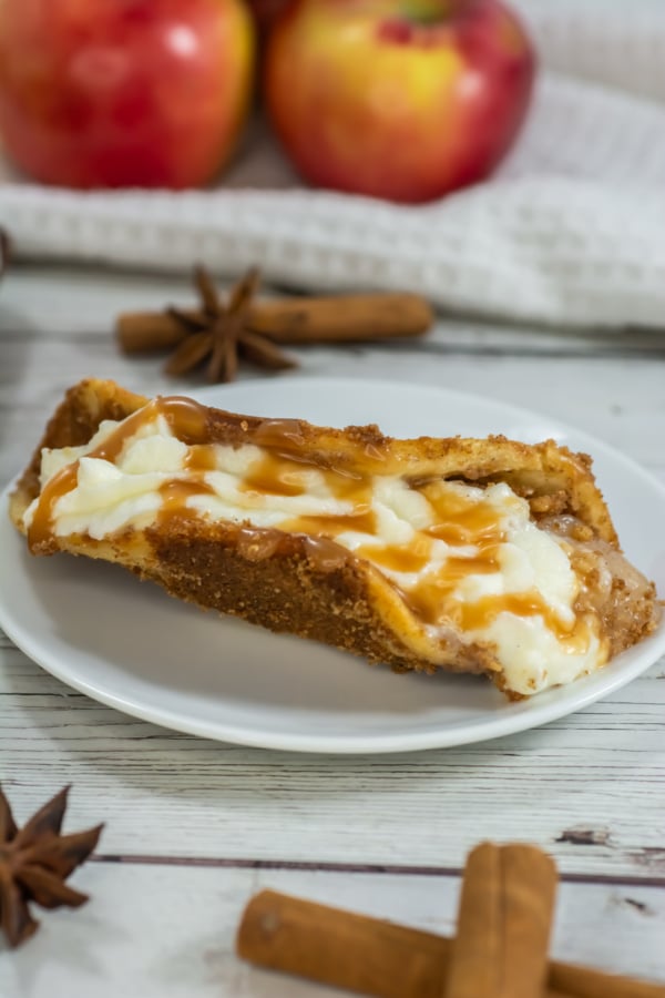 A slice of apple cheesecake topped with caramel sauce on a white plate, with apples, cinnamon sticks, and star anise in the background.