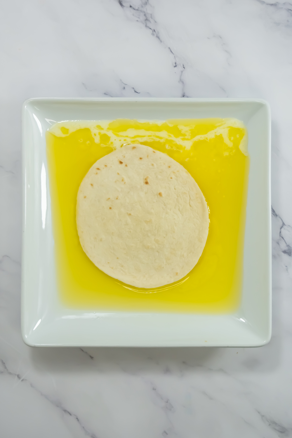 A round tortilla sits on a white square plate partially covered in melted yellow butter, on a marble surface.