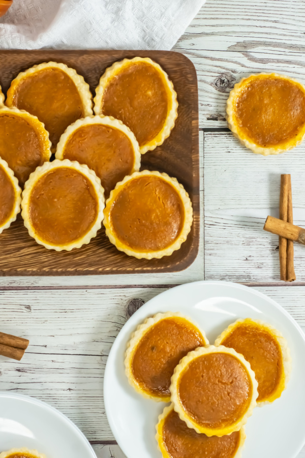 Mini pumpkin pies arranged on a wooden board and white plates, with two cinnamon sticks on a white wooden surface.