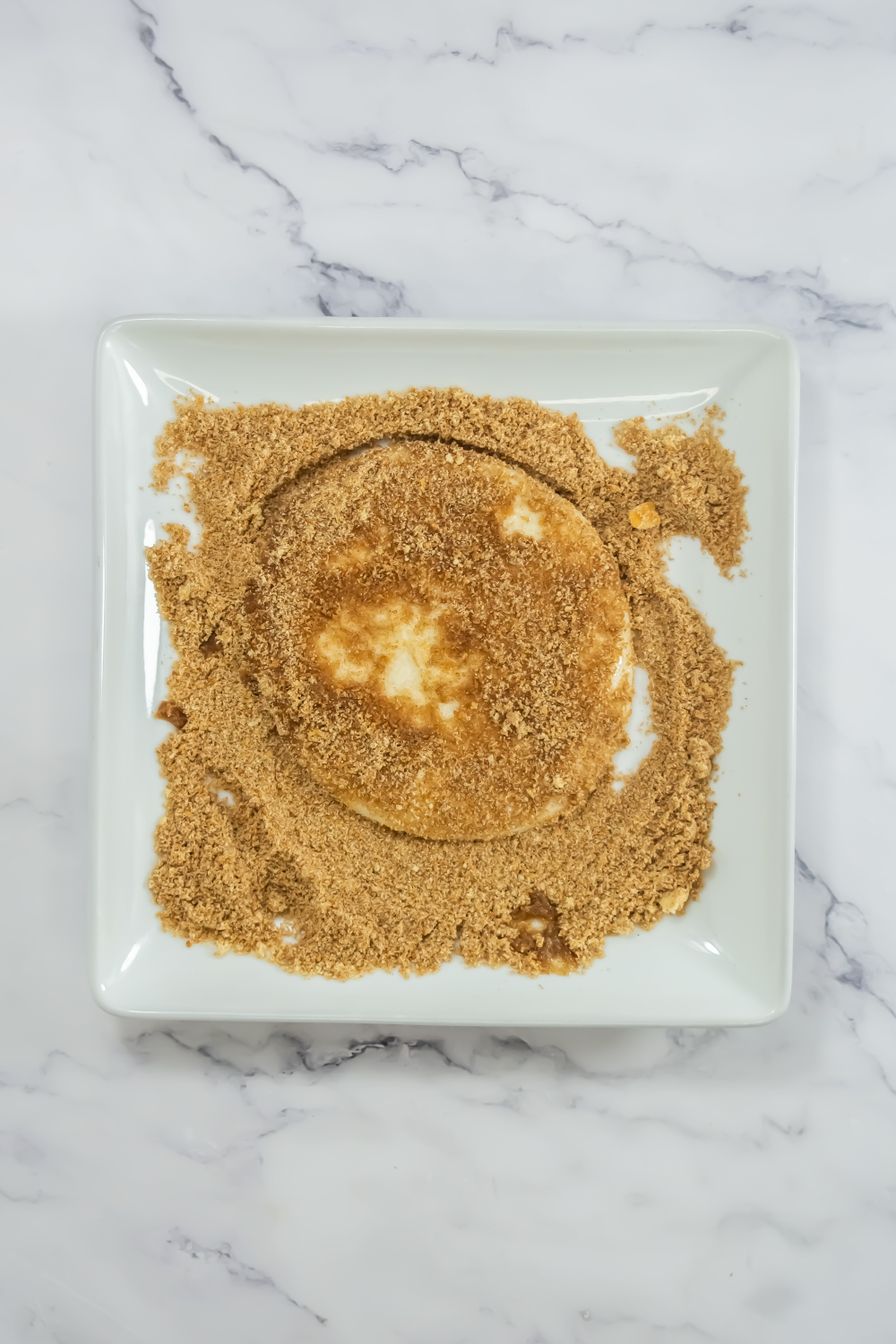 A round piece of bread coated with a layer of brown sugar sits on a white square plate, on a marble surface.