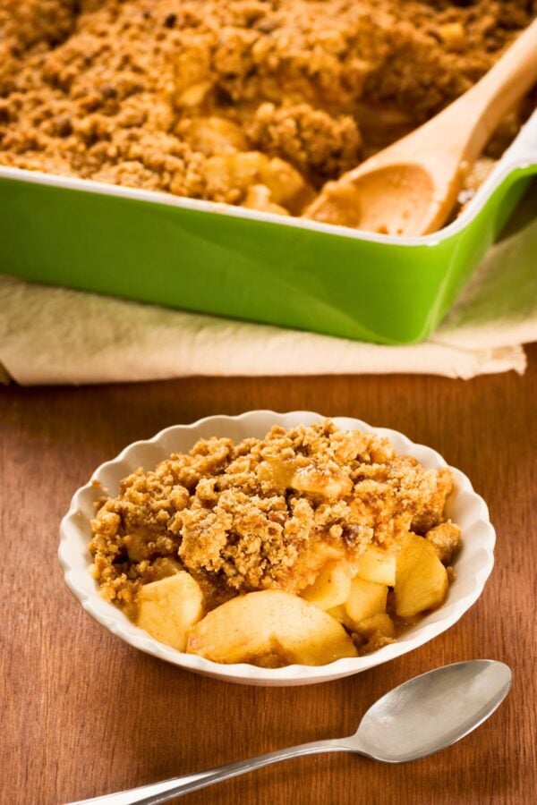 A bowl of apple crisp with a crumbly topping sits on a wooden table, with a spoon beside it and a baking dish of apple crisp in the background.