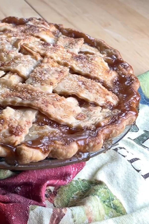 A close-up of a baked pie with a golden, lattice crust and sugar sprinkled on top, with some filling bubbling over the edge. The pie sits on a patterned cloth.