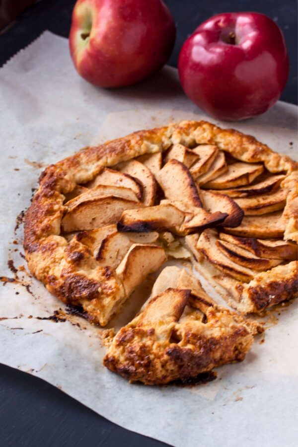 A rustic apple galette with a slice missing sits on parchment paper, with two whole red apples in the background.