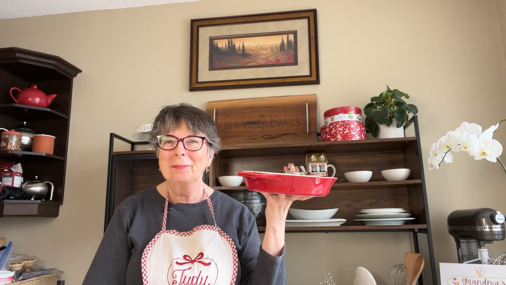 Older woman with glasses and short gray hair holds a red baking dish in a kitchen, wearing an apron that says "Truly Blessed." Shelves with dishes and decor are in the background.