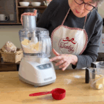 A person wearing an apron uses a food processor to mix butter, with a red measuring cup and ingredients on the wooden table.
