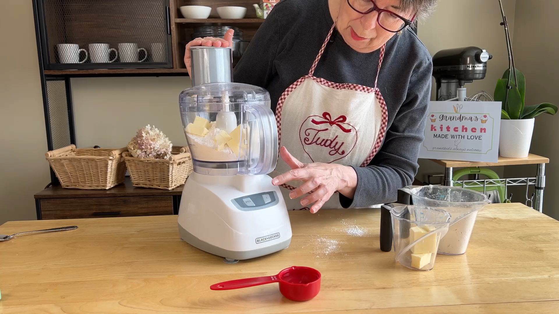 A person wearing an apron uses a food processor to mix butter, with a red measuring cup and ingredients on the wooden table.