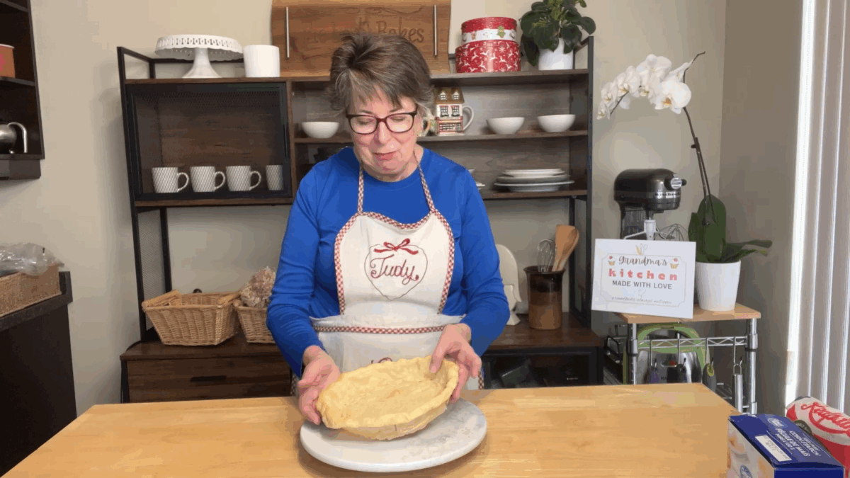 A woman wearing glasses and a blue shirt stands in a kitchen, holding an unbaked pie crust on a plate in front of her.