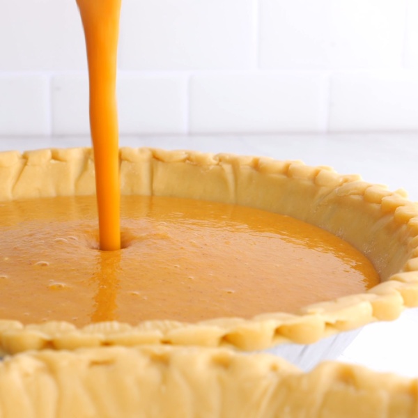 Pumpkin pie filling being poured into an unbaked pie crust on a white countertop with a white tile background.