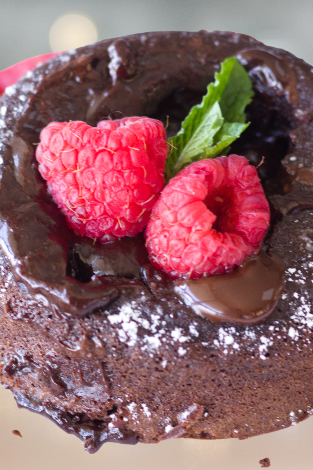 A close-up of a chocolate lava cake topped with two raspberries, a sprig of mint, and a dusting of powdered sugar. Melted chocolate is visible on top.