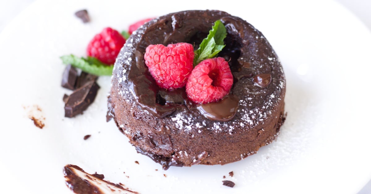 A gooey chocolate lava cake topped with raspberries and a mint leaf, served with melted chocolate and powdered sugar on a white plate.