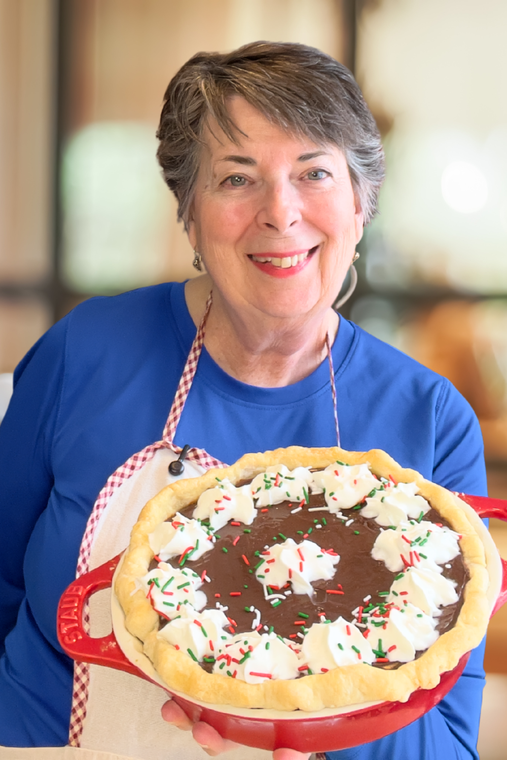 An older woman wearing a blue shirt and apron smiles while holding a chocolate cream pie topped with whipped cream and colorful sprinkles.
