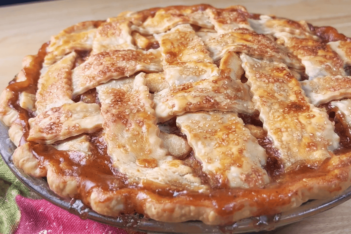 A baked pie with a golden brown lattice crust sits on a wooden surface. Sugar crystals are visible on top of the crust.
