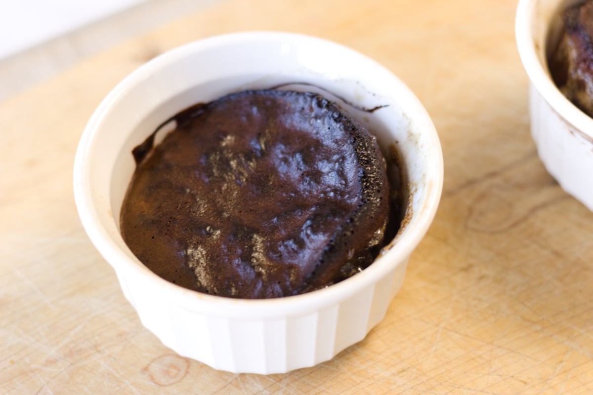 A baked chocolate dessert in a white ramekin sits on a wooden surface.