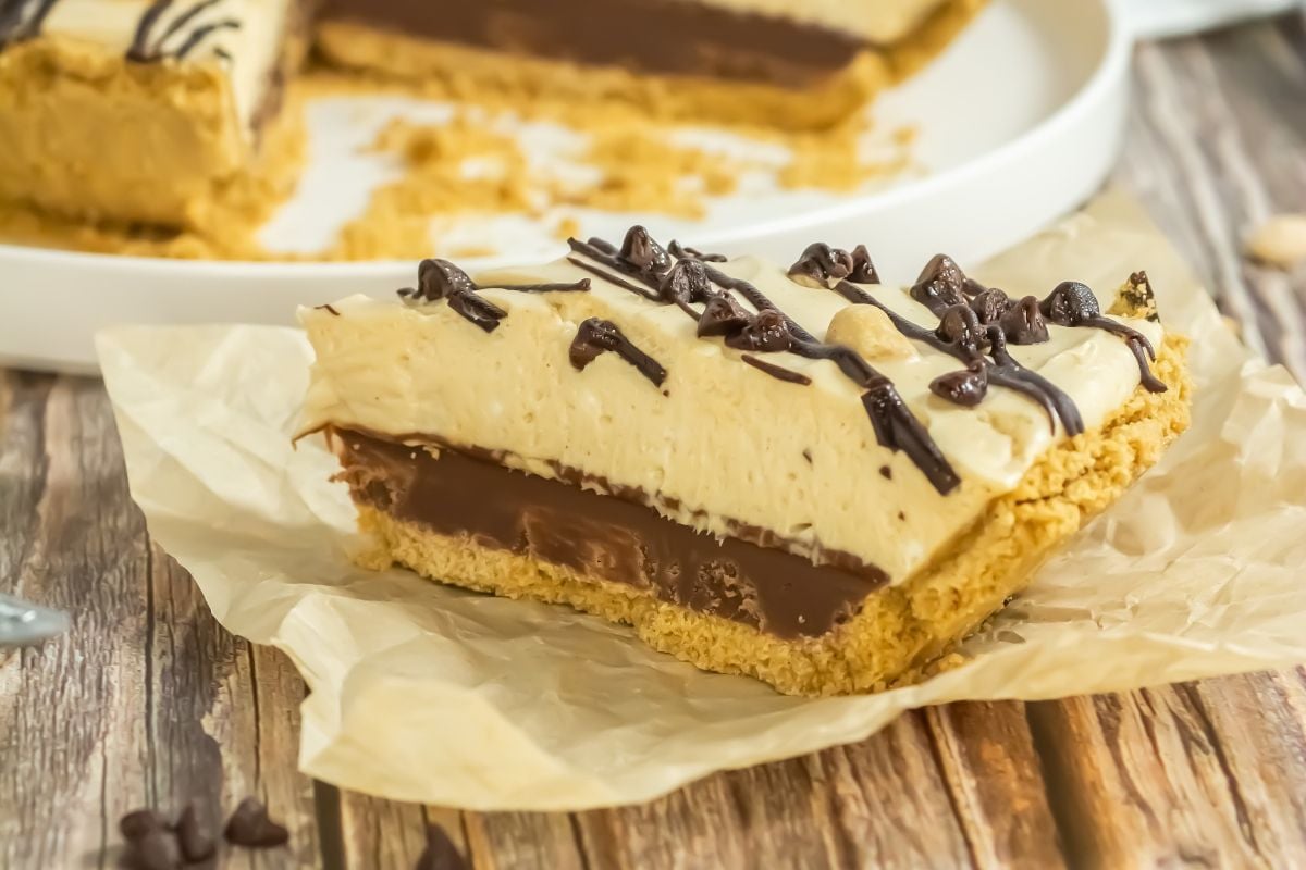 A slice of layered chocolate and cream pie with a graham cracker crust, topped with chocolate chips and drizzle, sits on parchment paper.