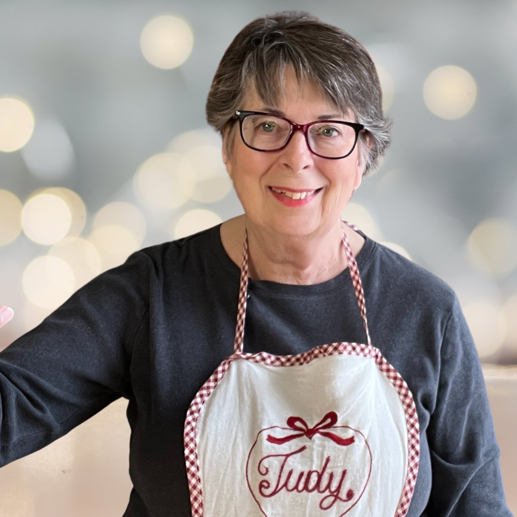 A woman with short gray hair and glasses wears a gray shirt and an apron that says "Judy," smiling and standing in front of a blurred background with lights.