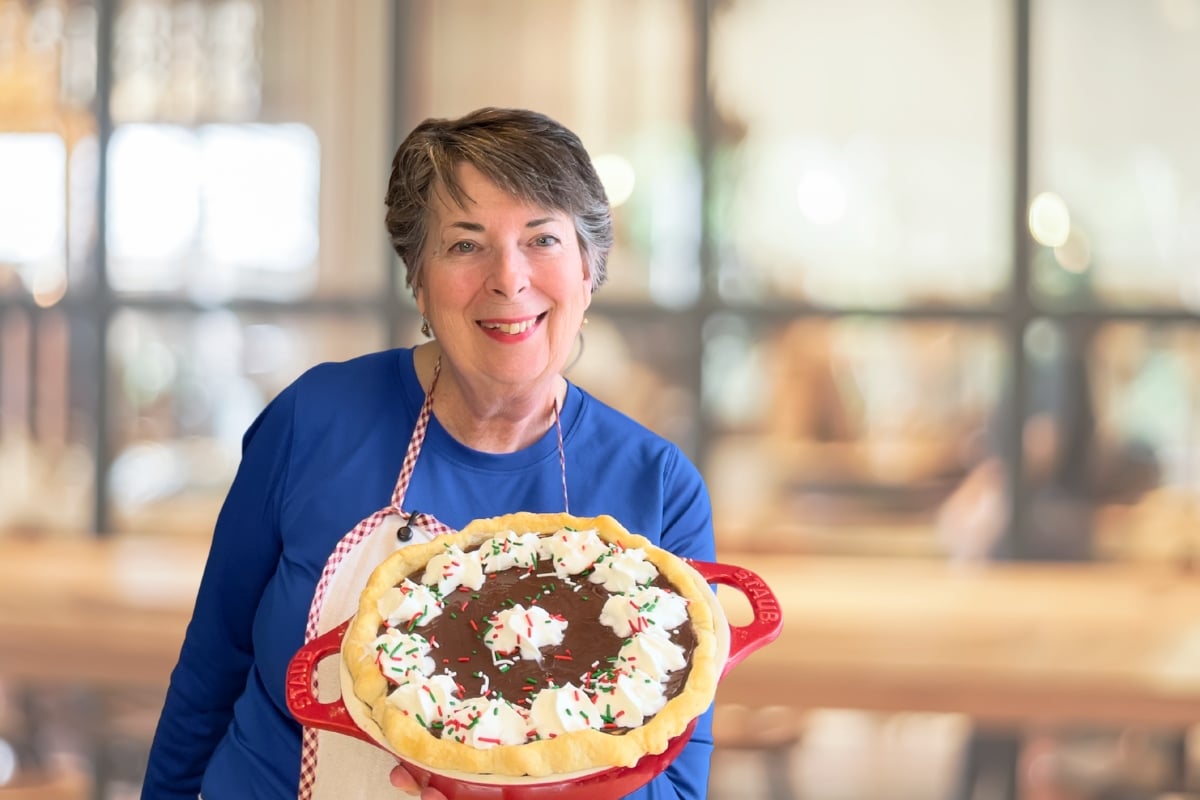 Older woman in a blue shirt holds a decorated chocolate pie with whipped cream and sprinkles, standing in a cozy, blurred kitchen or café setting.