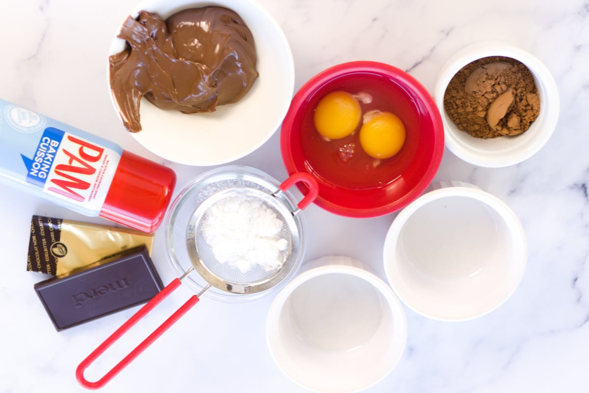 Bowls containing chocolate spread, cocoa powder, eggs, and baking chocolate, with a can of nonstick spray, powdered sugar in a sifter, and two empty ramekins on a marble surface.