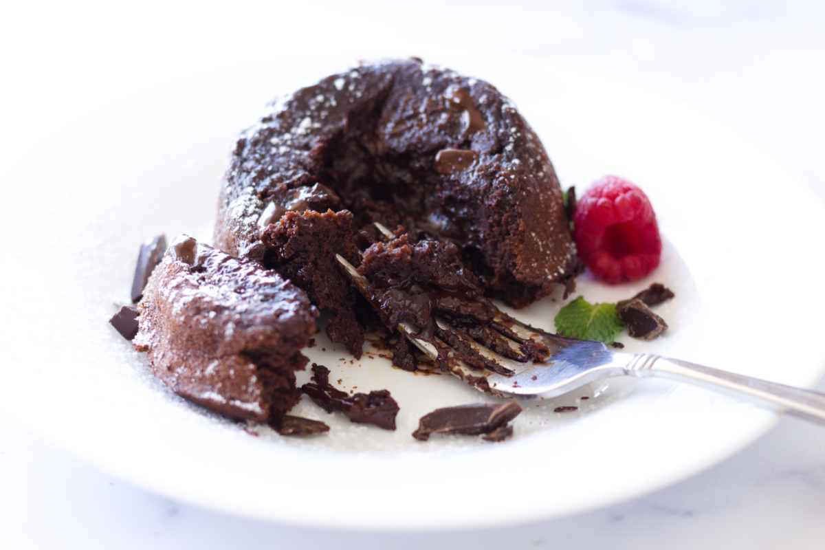 A partially eaten chocolate lava cake with molten center is on a white plate, garnished with a raspberry, mint leaf, chocolate pieces, and a fork.