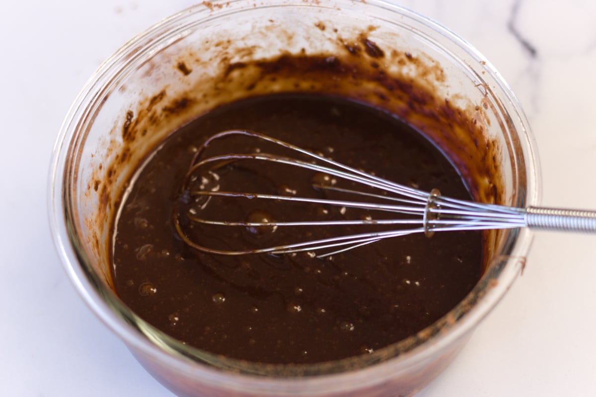 A glass bowl containing chocolate batter being mixed with a metal whisk, sitting on a white surface.