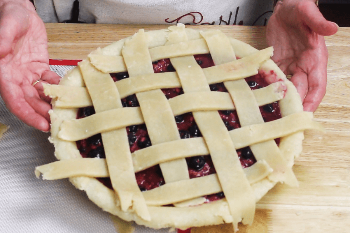 A person holds an unbaked berry pie with a lattice crust on a wooden surface.
