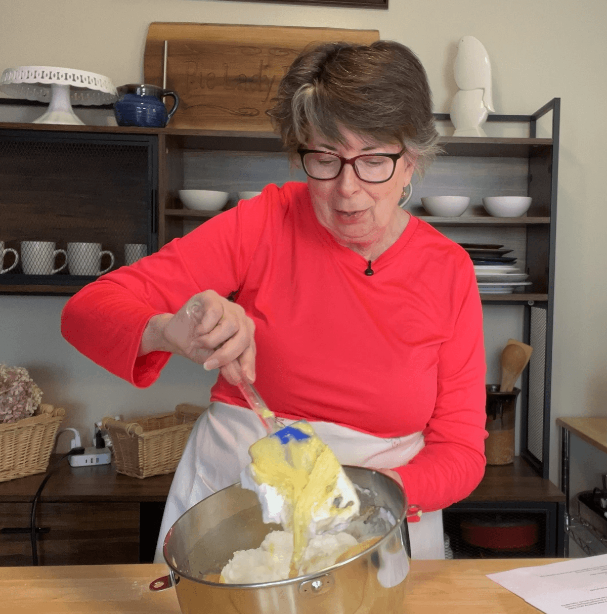 A woman in a bright red shirt mixes ingredients in a metal bowl with a spatula in a kitchen setting.