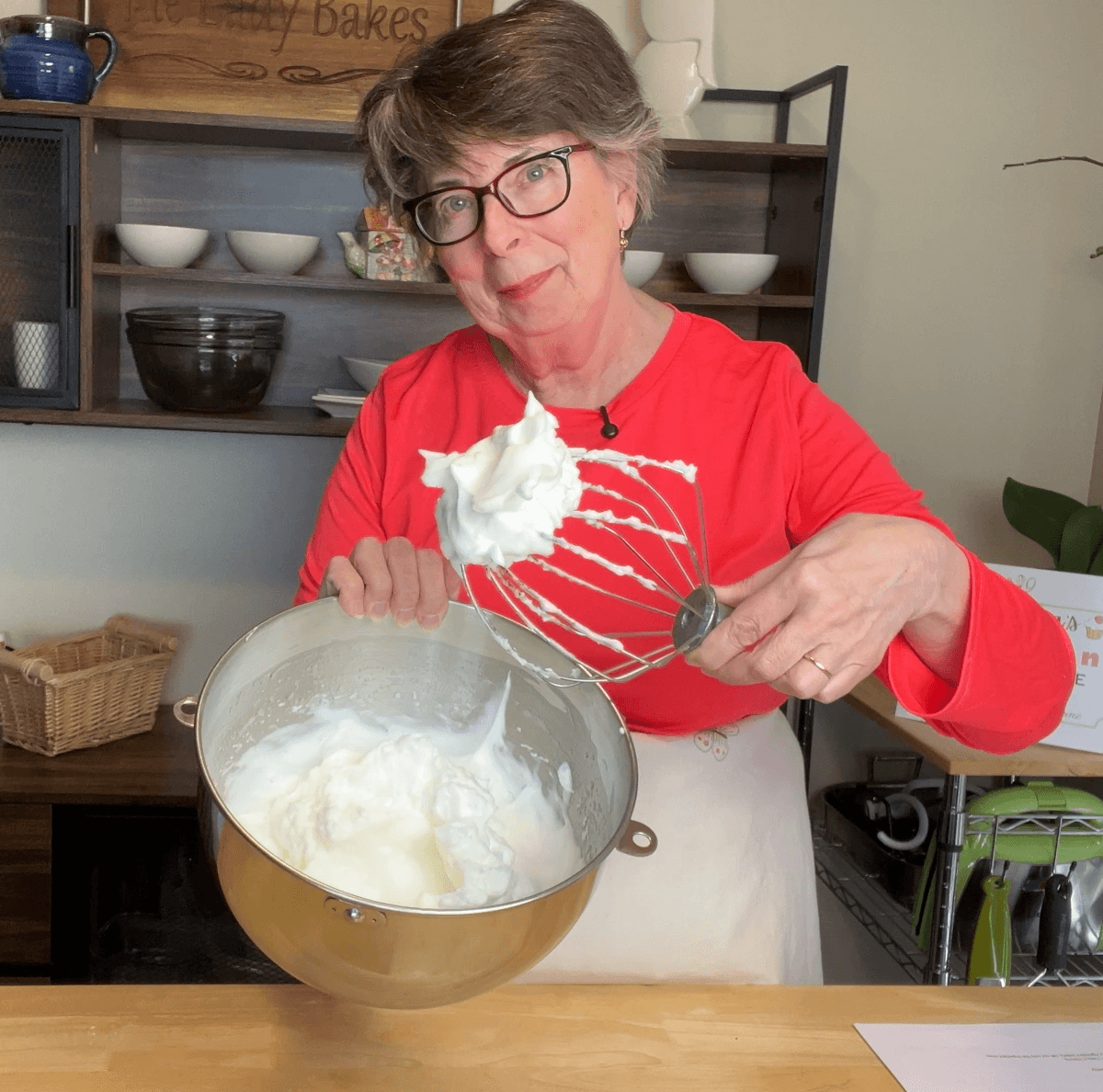 A person in a red shirt holds a mixing bowl of whipped egg whites and a whisk covered in the mixture, standing in a kitchen.