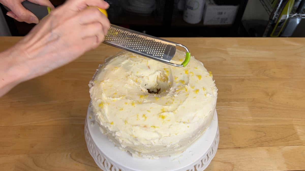 A person grates lemon zest over a frosted bundt cake on a white cake stand, positioned on a wooden countertop.